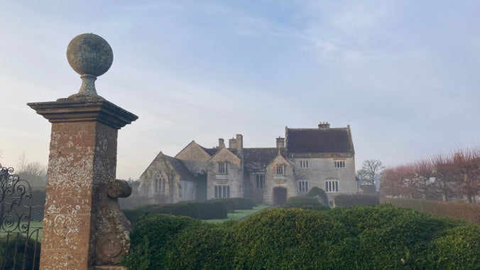 A stone mansion set back behind stone gate posts and a formal garden